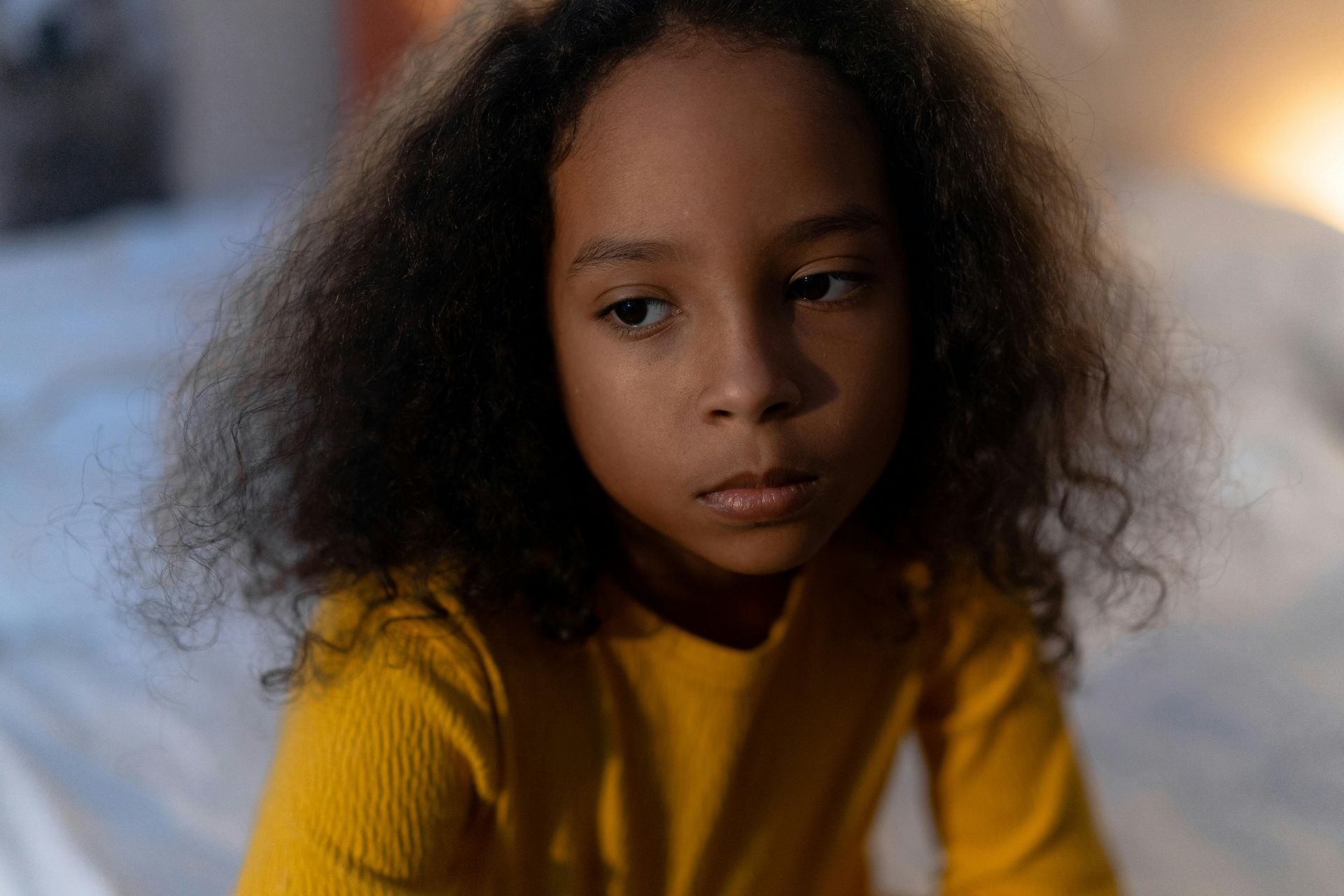 Girl in yellow shirt looking somber, sitting on a bed.