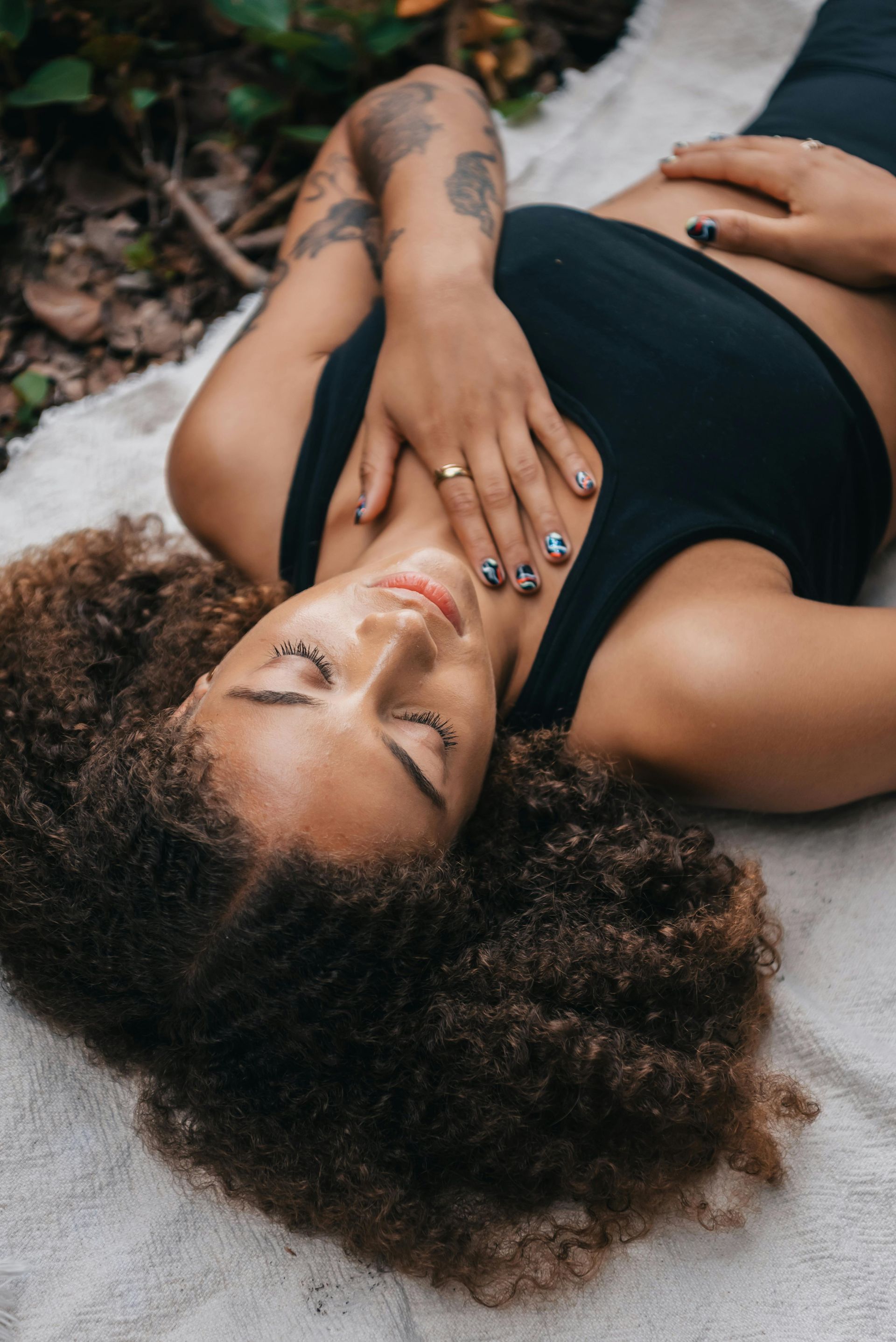 Woman with curly hair reclines outdoors, eyes closed, hands on chest and abdomen, wearing a black top.