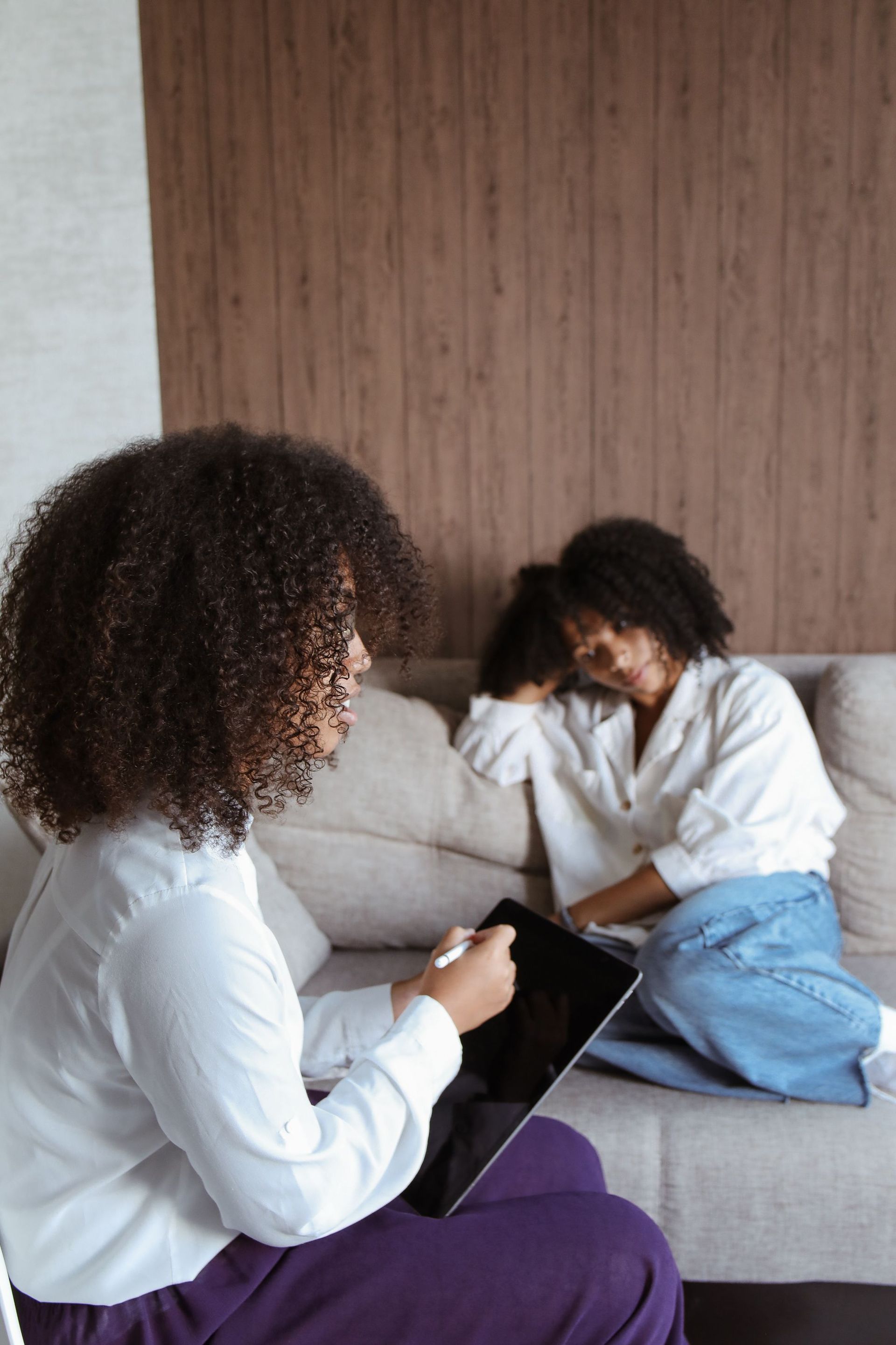 Two African American women. Black therapist supporting black client. Client is seated on sofa. Therapist providing client with support.