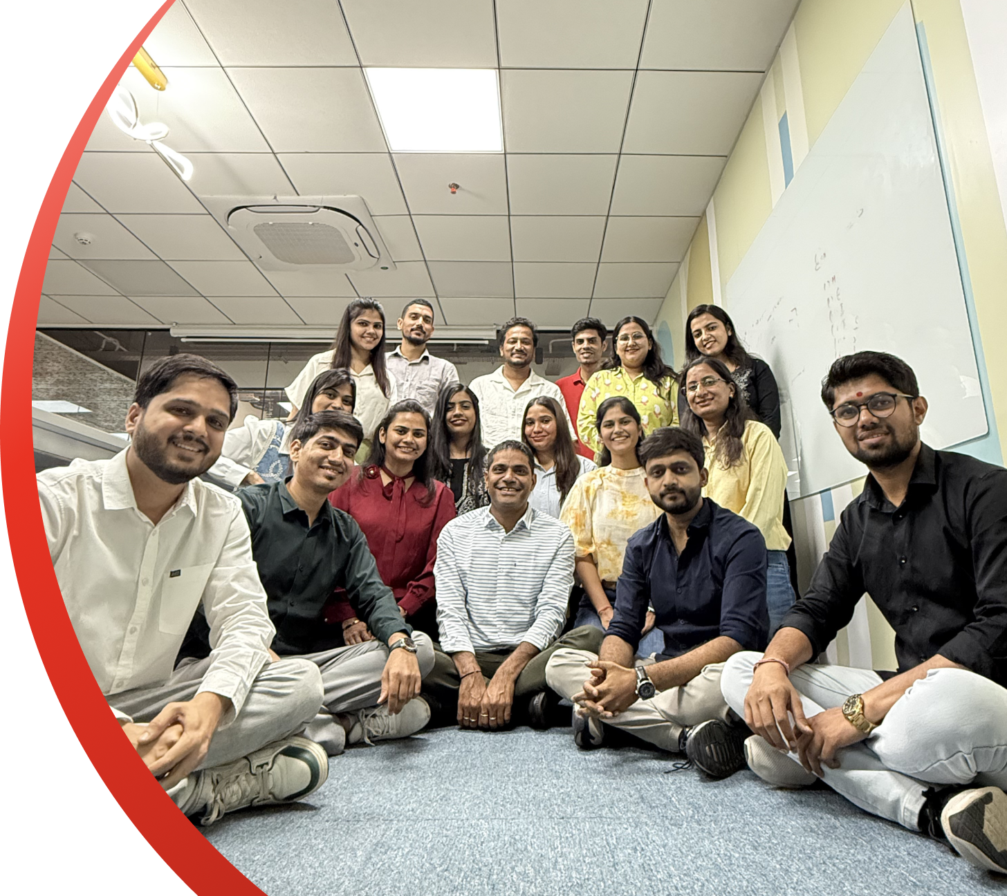 Group of coworkers posing on a carpeted office floor in a bright hallway, smiling at the camera.