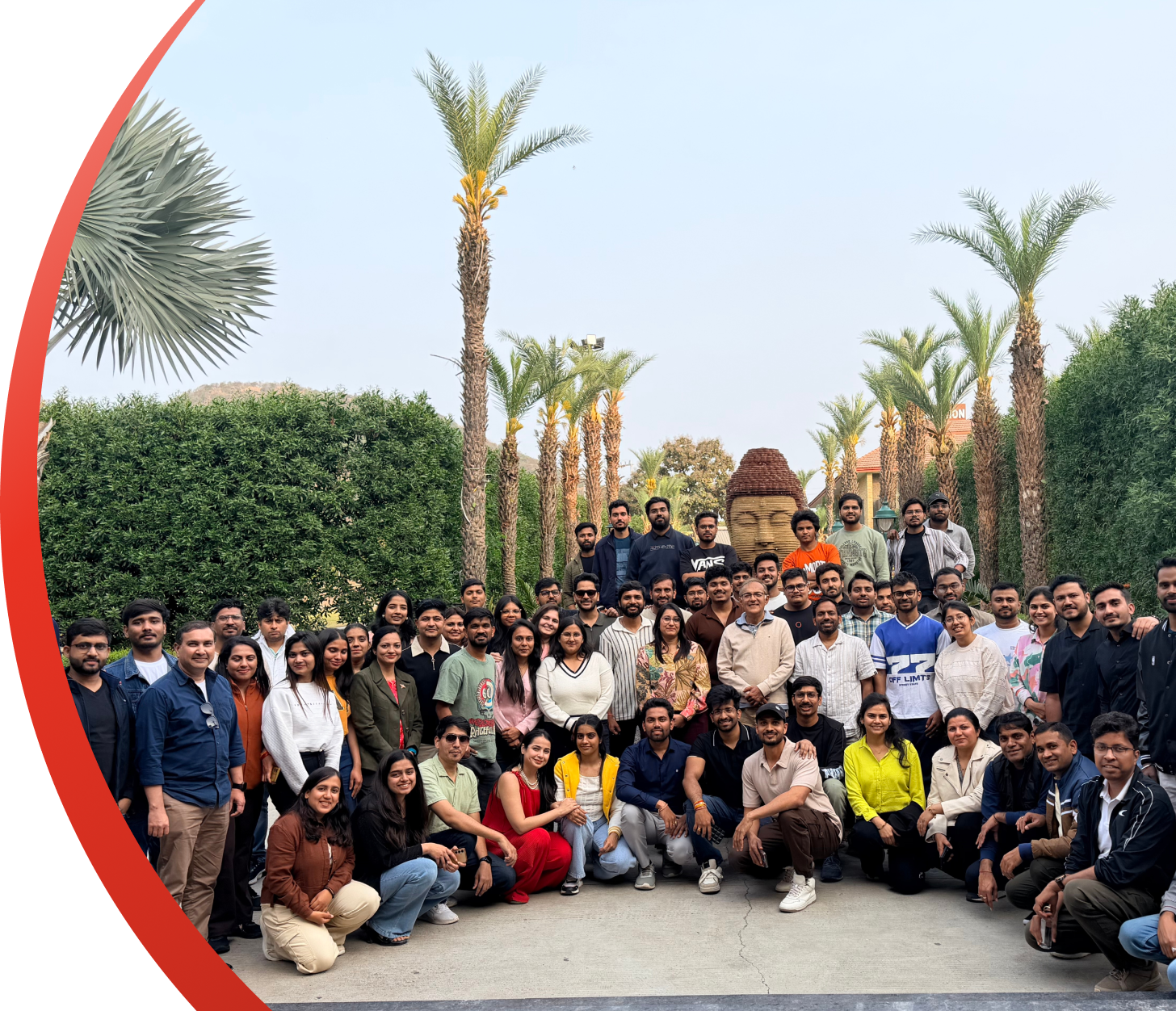 Large group posing outdoors in front of palm trees and hedges, with a statue centered behind them