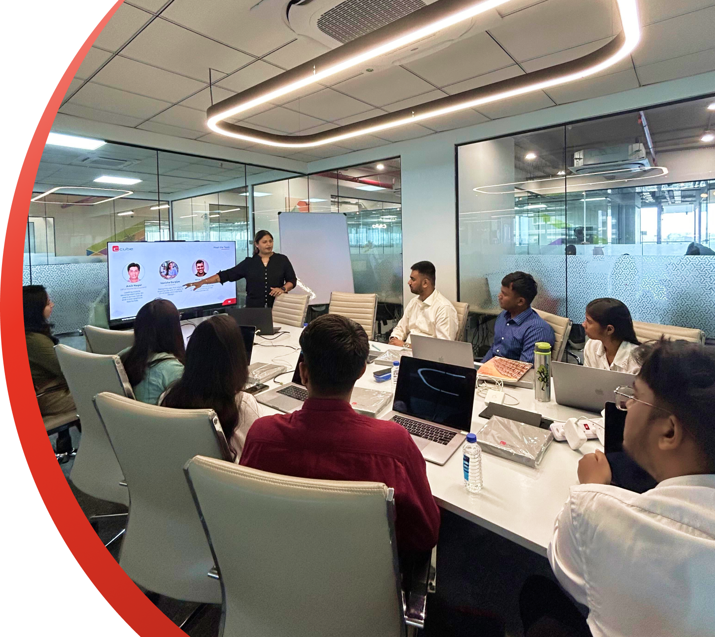Team meeting in a modern conference room, with a presenter speaking beside a screen and attendees seated around a table.