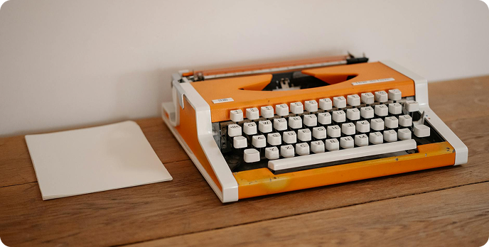 Orange typewriter on a wooden desk beside a blank sheet of paper