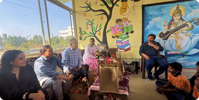 People seated outdoors in a colorful courtyard beside murals and a tree, with one person kneeling in front.