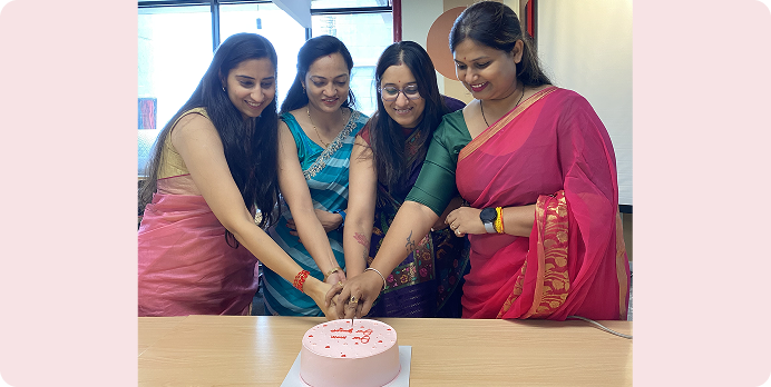 Four women in colorful dresses cut a pink cake together at a celebration table.