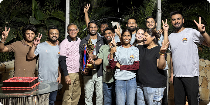 Group of people posing outdoors at night around a birthday cake, smiling and celebrating.