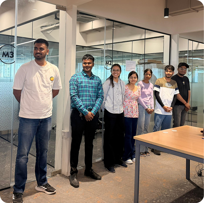 Group of people standing in an office hallway beside glass-walled rooms and a table