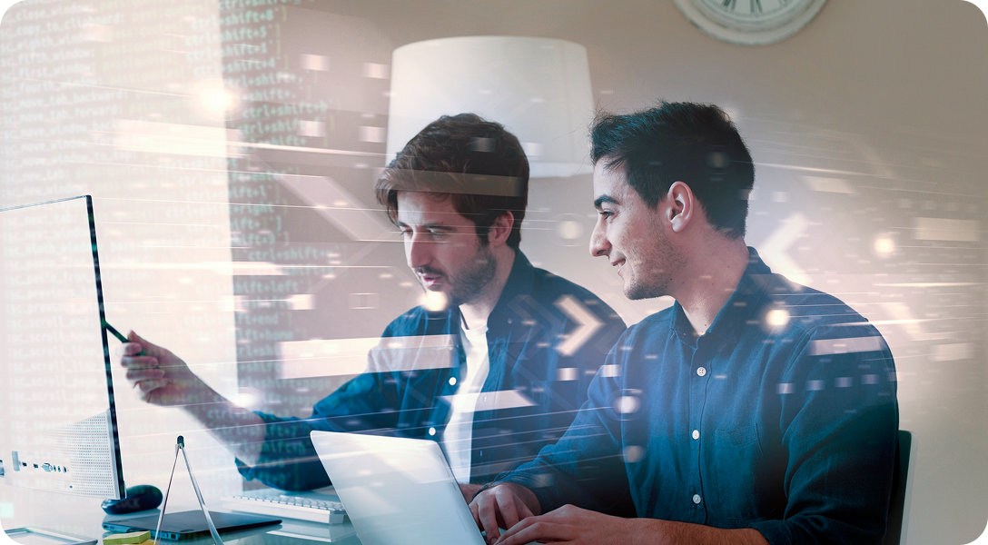 Two coworkers reviewing data on a laptop and monitor in a bright office, one pointing at the screen.