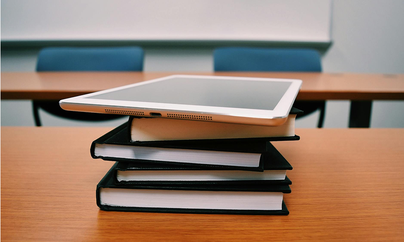 Stack of books with a tablet on top on a wooden table in a classroom or study room.