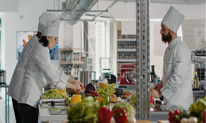 Two chefs in white uniforms prepare vegetables in a bright commercial kitchen