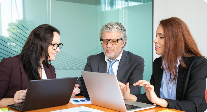 Three colleagues in business attire discuss documents and laptops in a glass-walled office