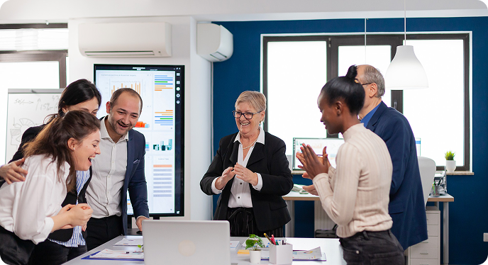 Coworkers in a modern office meeting around a laptop, discussing charts on wall screens