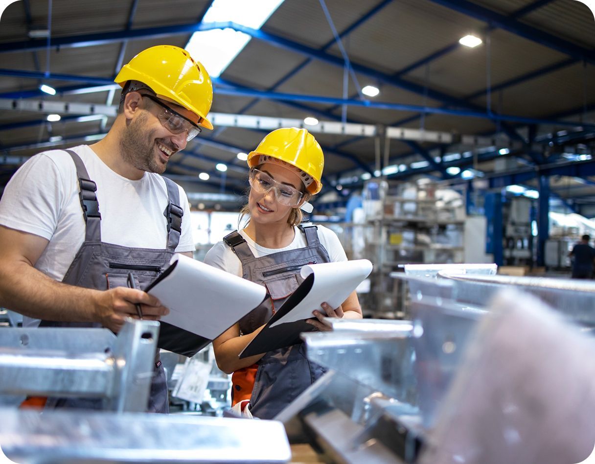 Two workers in yellow hard hats reviewing paperwork on a factory floor.