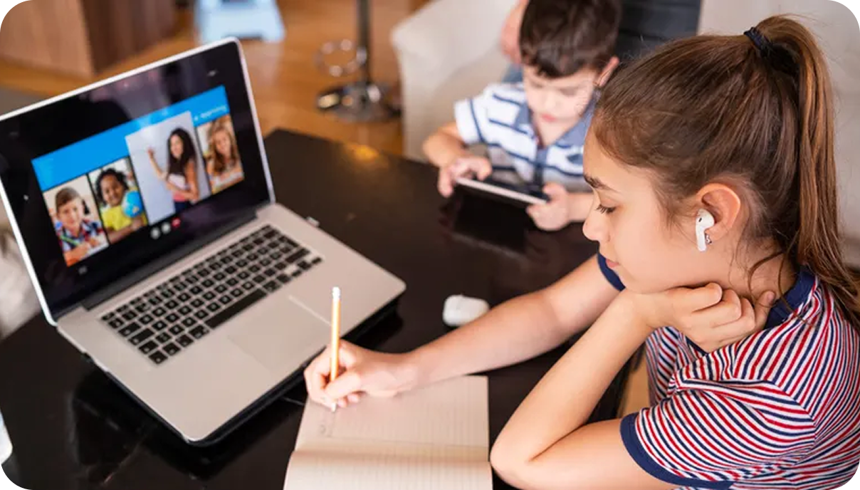 Two children on a video call, one writing in a notebook beside a laptop screen showing participants.