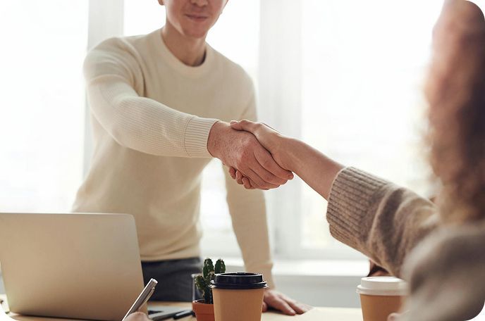 Two people shaking hands across a table with a laptop and coffee cups in a bright office
