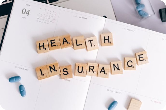 Scrabble tiles spelling “HEALTH INSURANCE” on paperwork with blue pills scattered nearby.