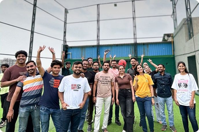 Group of people posing and smiling on a grassy sports field, some waving under a netted enclosure.