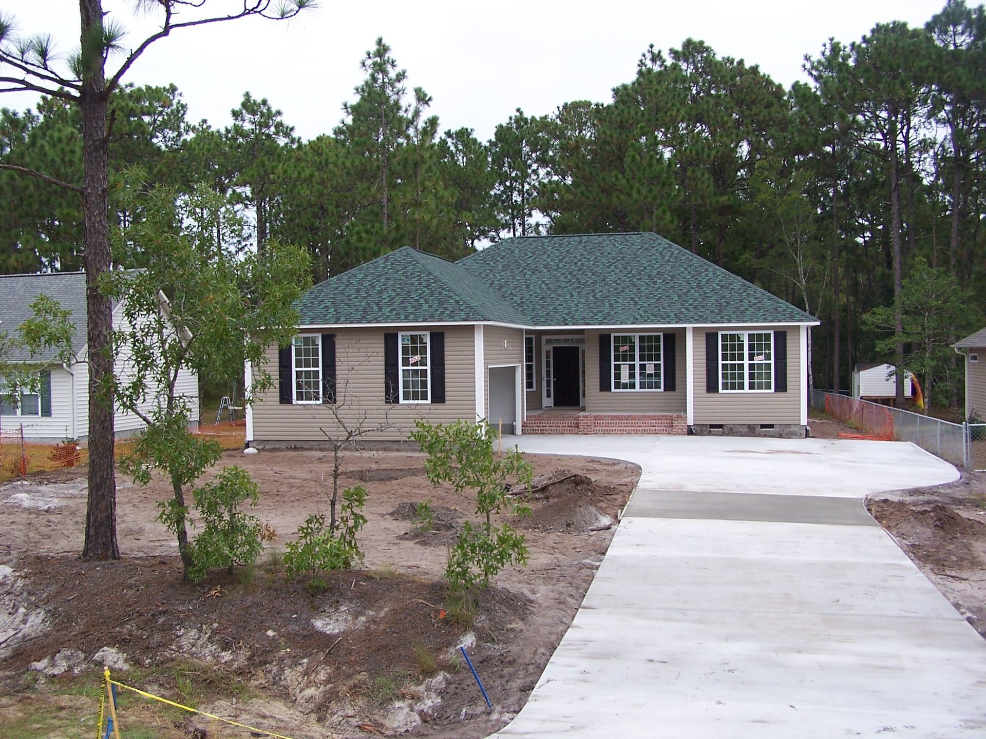 New single-story house with green roof, concrete driveway, and surrounding trees.