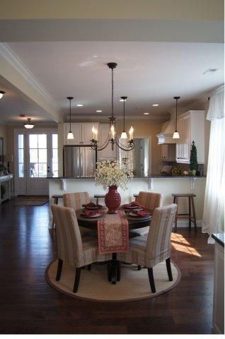 Dining room with round table, chairs, chandelier, and a vase of flowers. Dark wood floor.