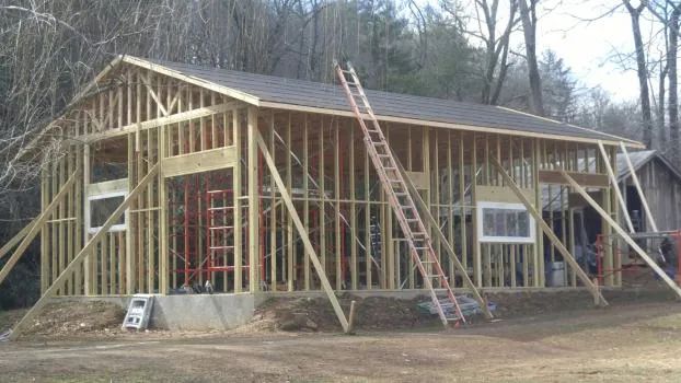 Wooden structure under construction, with roof and support beams. A ladder leans against the building.