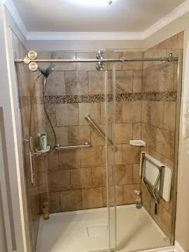 Accessible shower with grab bars and fold-down seat, beige tile, glass door.