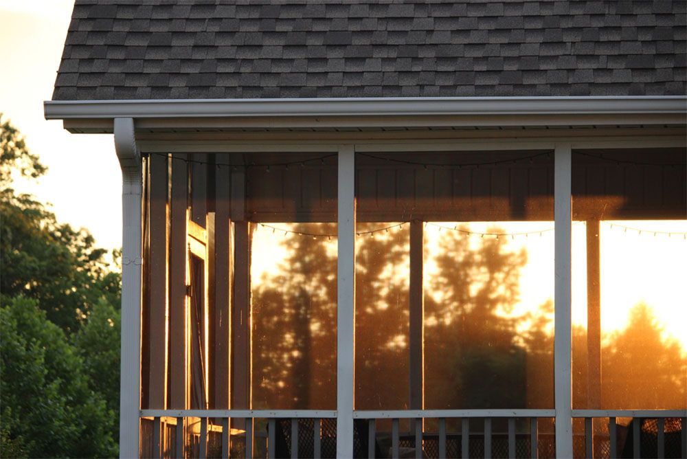 Sunset glowing through screened porch windows of a house with white railing and dark roof