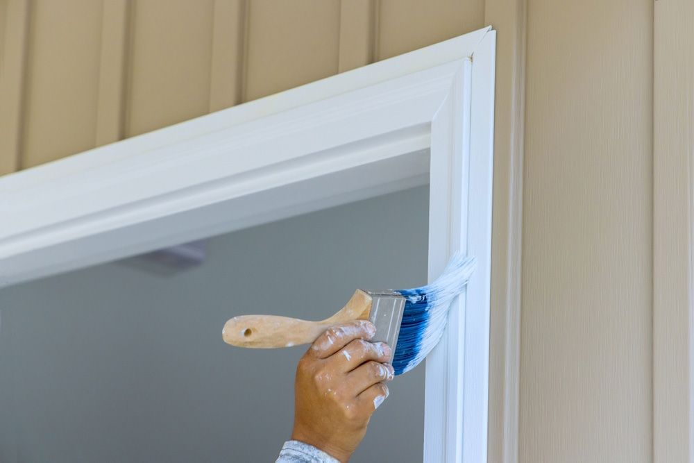A hand paints white trim around a doorway with a blue brush.