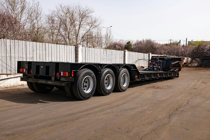 Black flatbed semi-trailer parked on a dirt lot beside a white fence and leafless trees