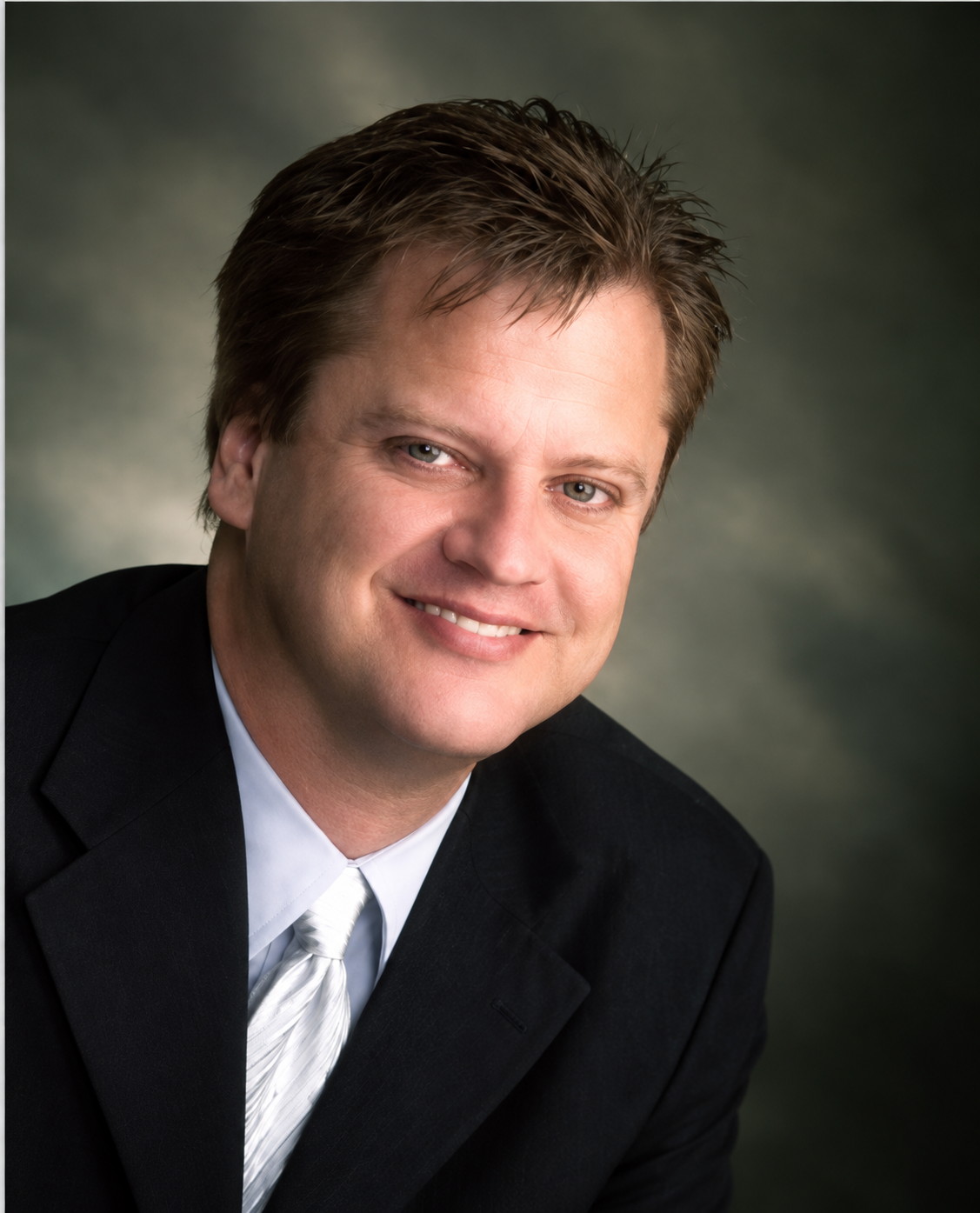 Studio headshot of a smiling person in a dark suit and tie against a gray background