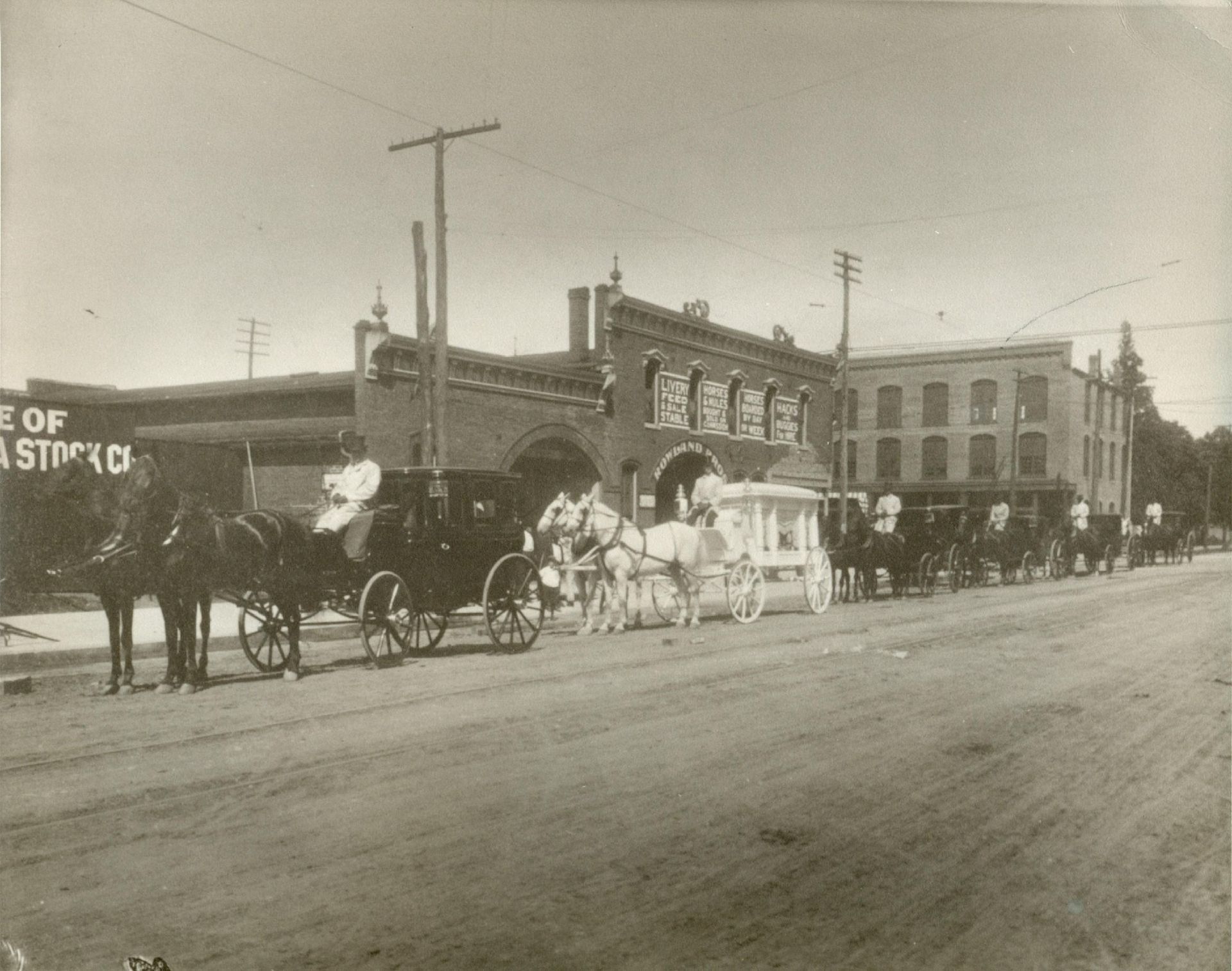 Horse-drawn carriages lined up on a dirt street beside brick storefronts in a historic town.