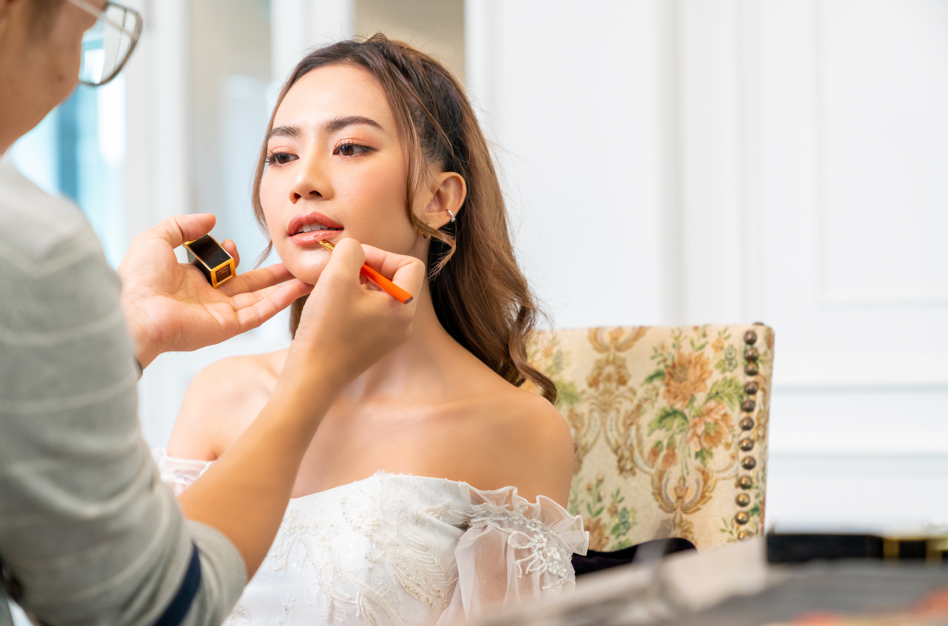 A woman in a white dress is getting her makeup done by a makeup artist.