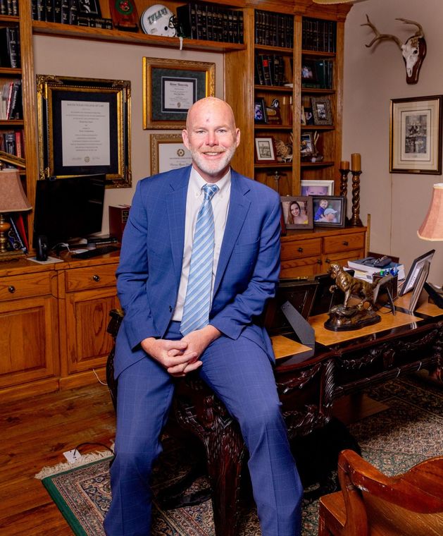 Man in blue suit, smiling, sits on desk in wood-paneled office.