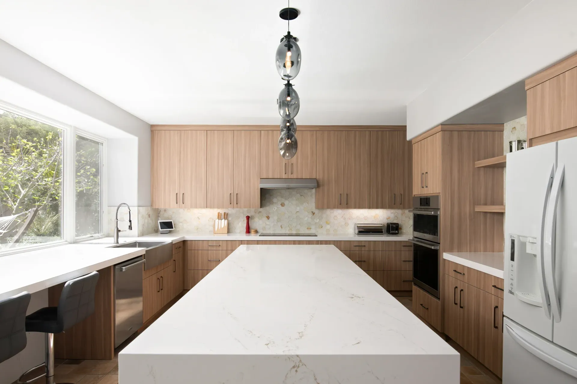Modern kitchen with white island, wood cabinets, stainless fridge, and large windows.