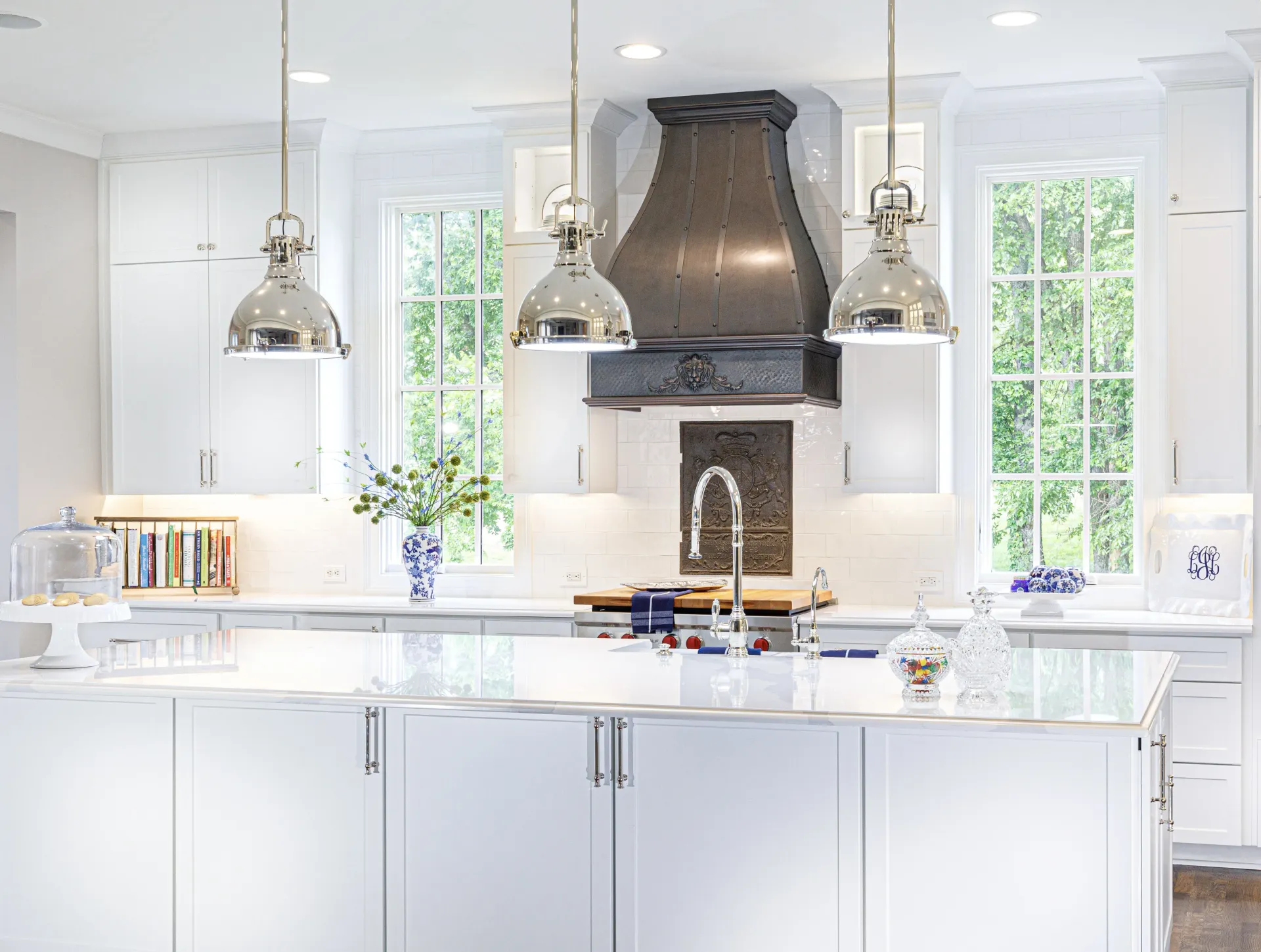 Bright white kitchen with marble island, hanging pendant lights, and large windows by a dark range hood