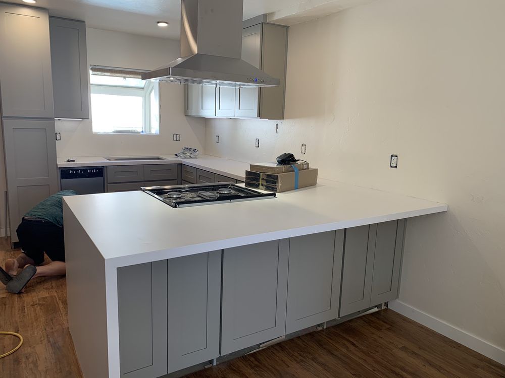 Modern kitchen with white island, gray cabinets, stainless hood, and wood floor; person crouched at left.