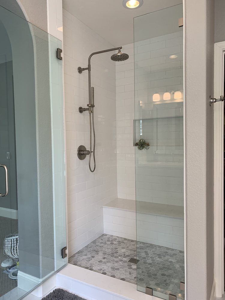 Glass shower stall with rainfall and handheld showerheads, white tile walls, and pebble floor.