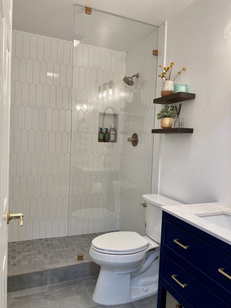 Small bathroom with glass shower, white tile, toilet, and dark blue vanity with shelves.