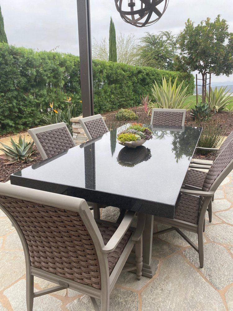 Outdoor patio dining table with six woven chairs, glossy black top, and potted plants in a garden setting