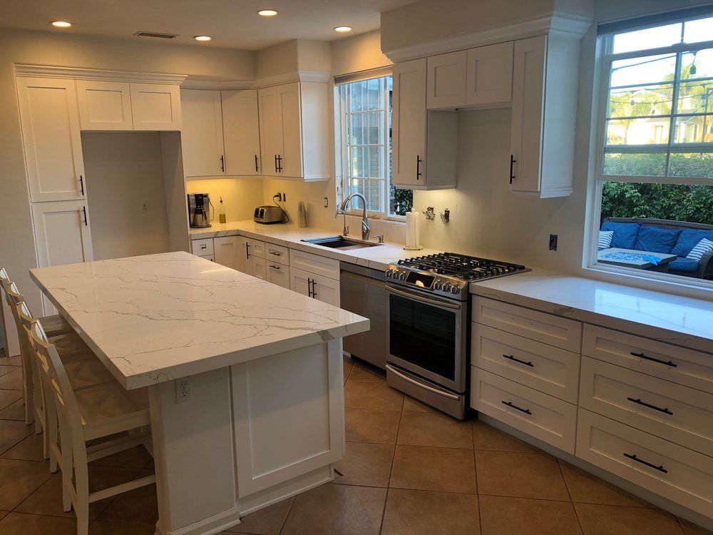 Bright white kitchen with large island, farmhouse sink, stove, and sunlit windows.