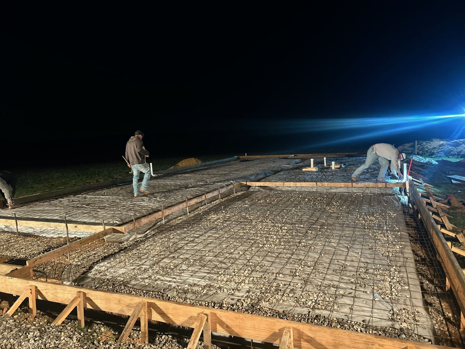 Two workers at night pour concrete for a large slab under bright floodlights.
