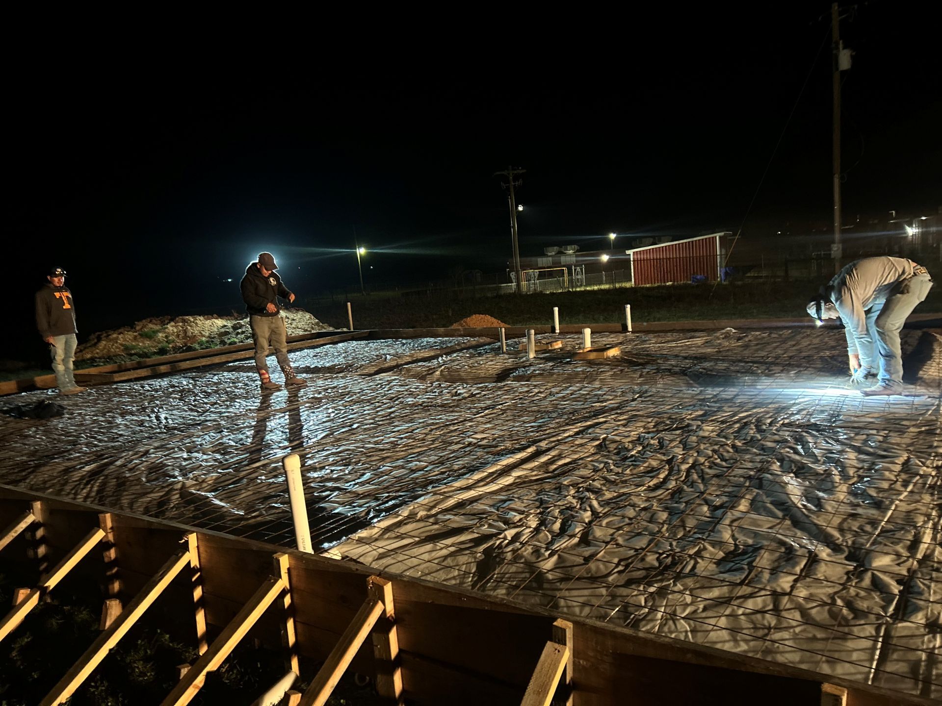 Workers on a snowy construction site at night, one shoveling under bright floodlights.