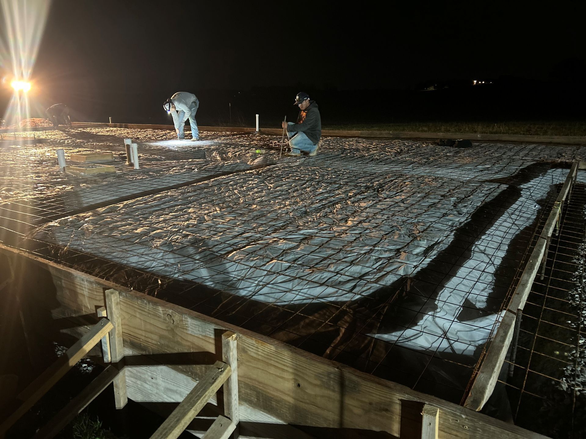 Workers laying rebar on a snow-dusted roof at night under bright work lights
