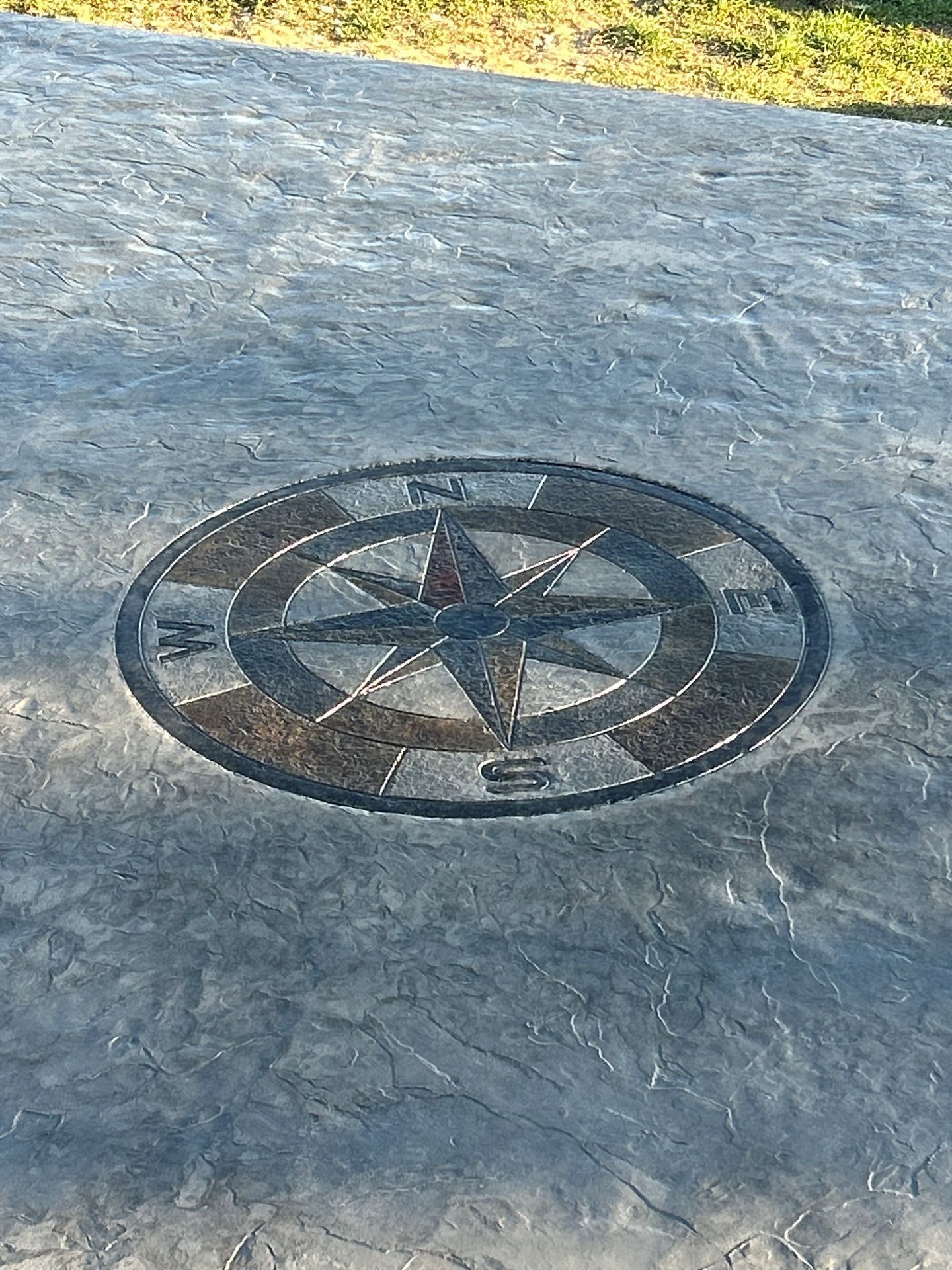 A compass rose mosaic embedded in a stone surface outdoors, with sunlight and grass at the top edge.