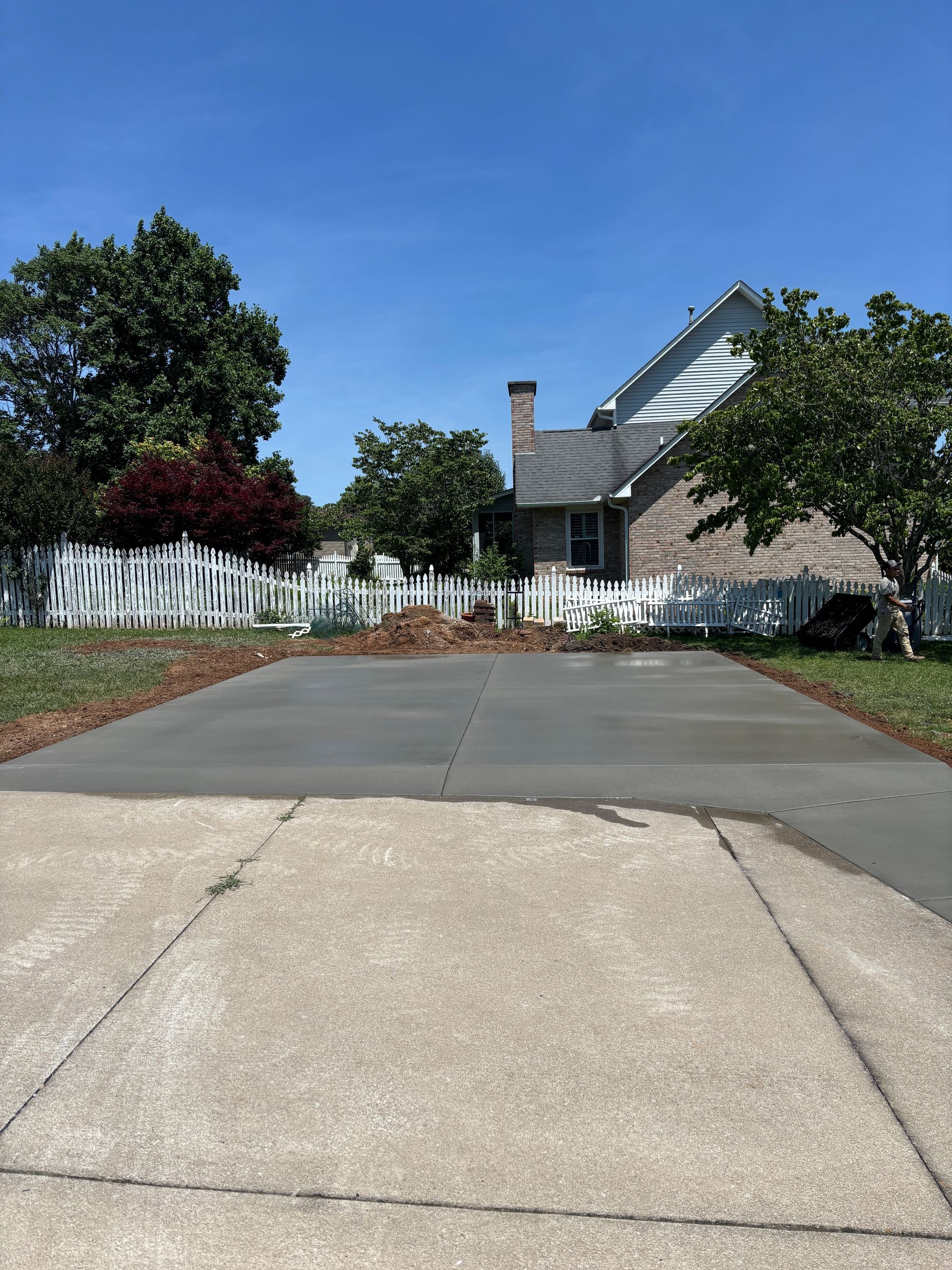 Residential driveway leading to a brick house with white fence and trees under a blue sky