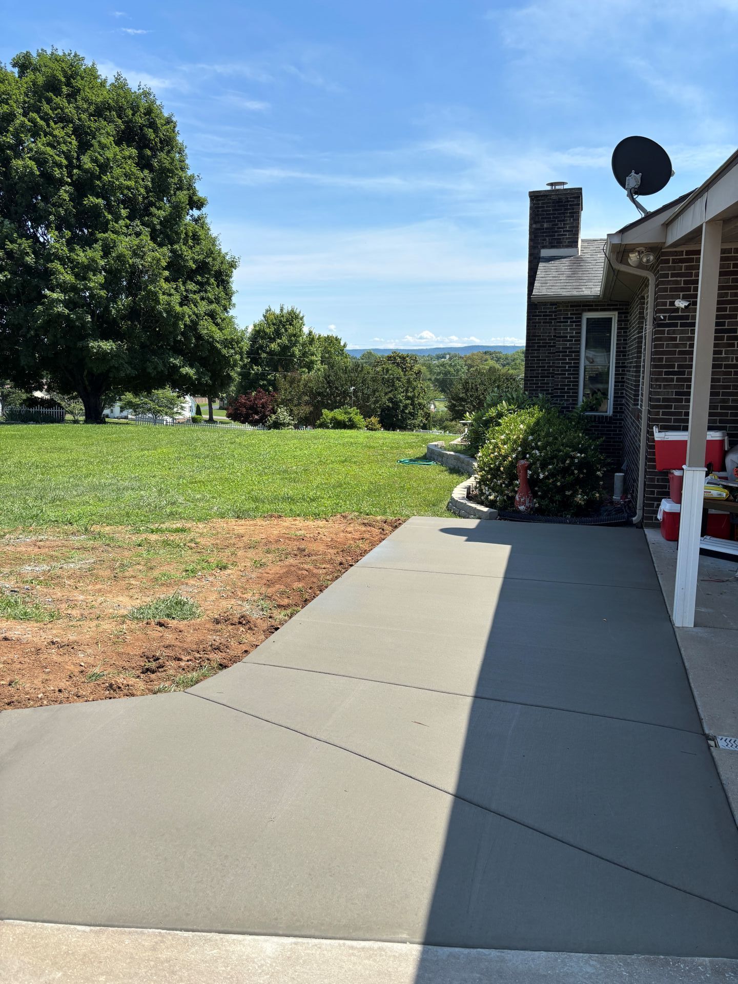 Concrete walkway beside a house, with a lawn, large tree, and patio furniture on a sunny day