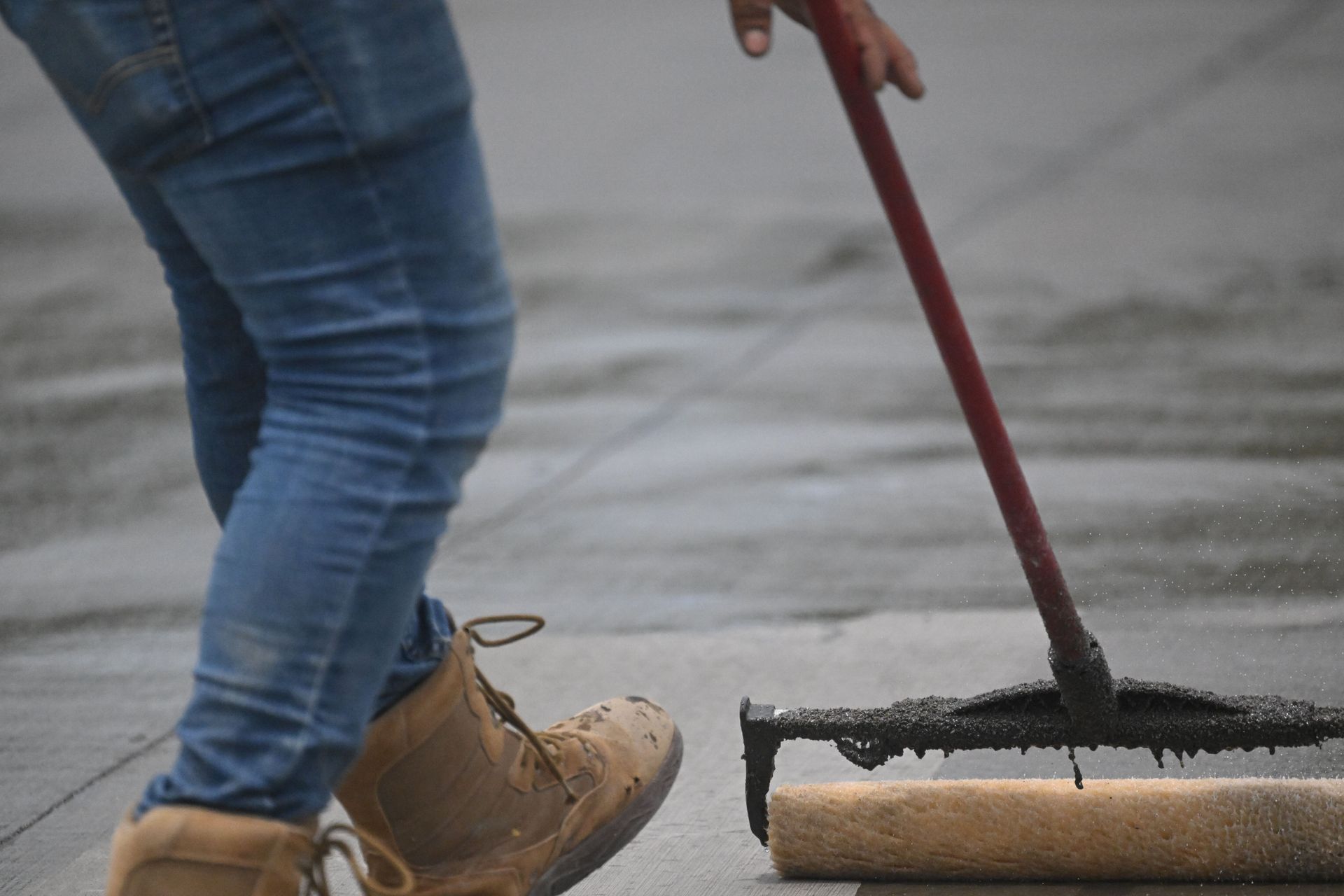 Worker pushing a squeegee on a wet pavement, wearing jeans and tan work boots.