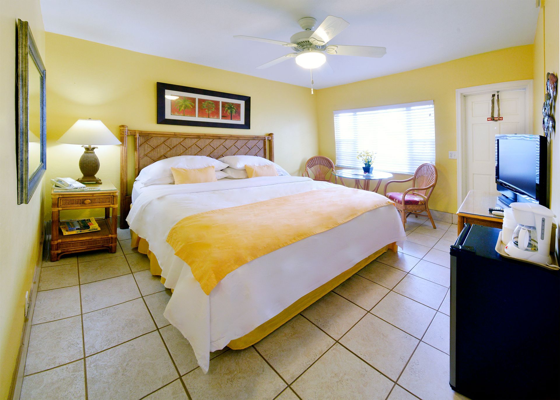 Yellow-walled hotel room with bed, table, chairs, and TV. Window, ceiling fan, and tiled floor.