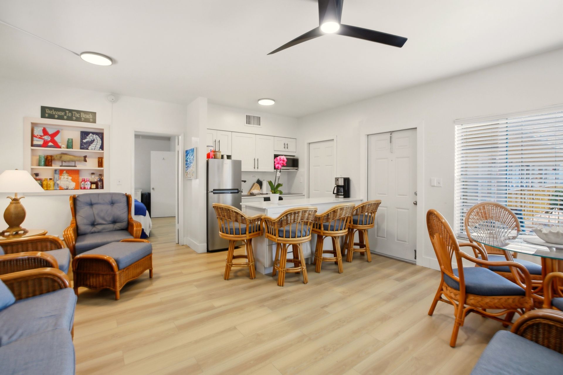 Bright open living-dining room with wicker chairs, blue armchairs, white walls, and a ceiling fan