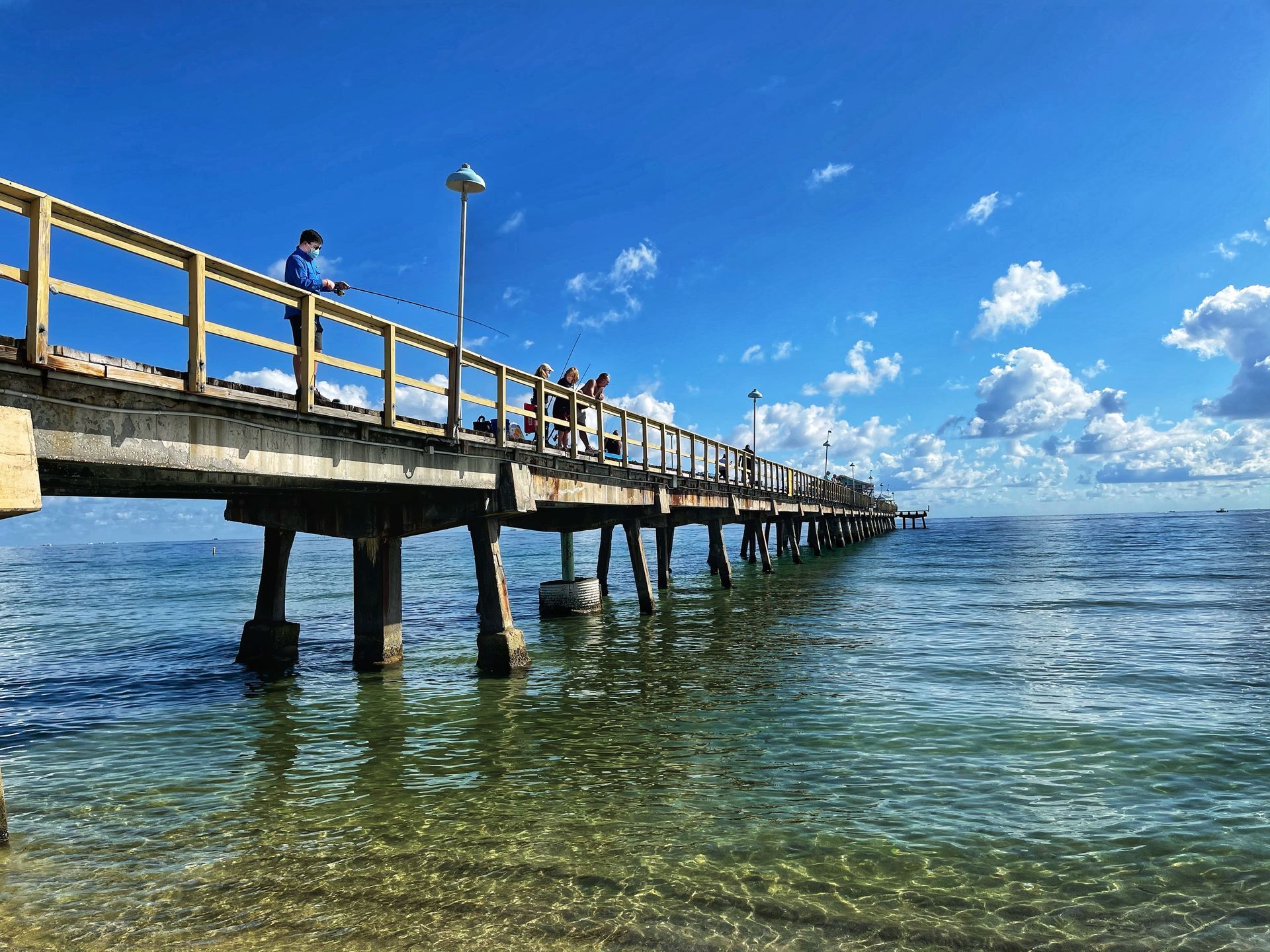Wooden pier extends into blue ocean, people fishing under a bright sky.