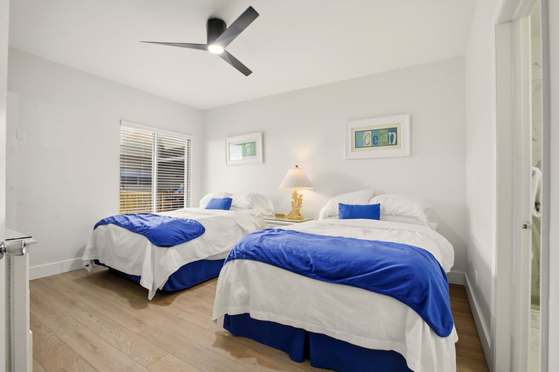 Bright bedroom with two twin beds, blue blankets, white walls, and a ceiling fan